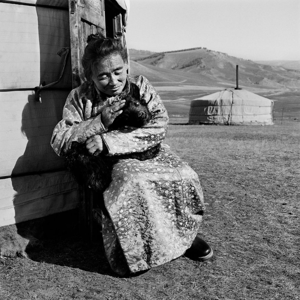 Black and white photo of an old woman looking tenderly at a small goat in her arms.