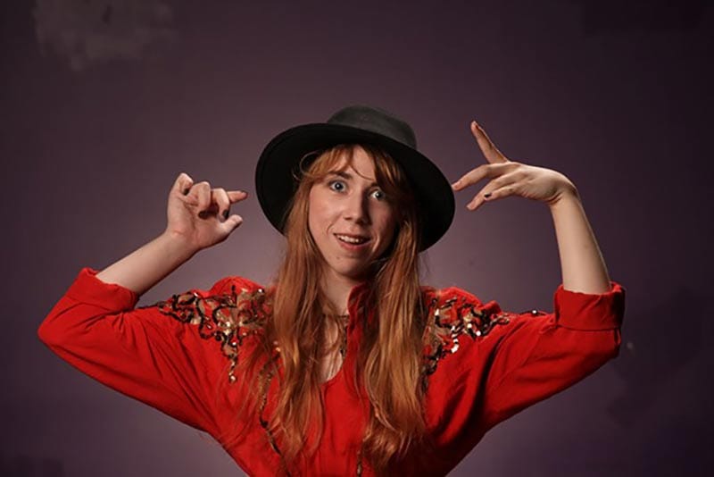 Tina Friml facing camera with both arms raised wearing a black fedora hat.