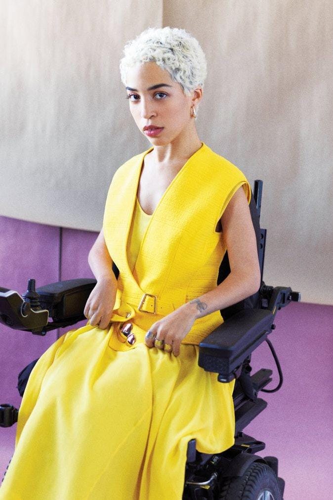 Jillian wears a yellow dress and lots of silver rings, sitting in a power chair.