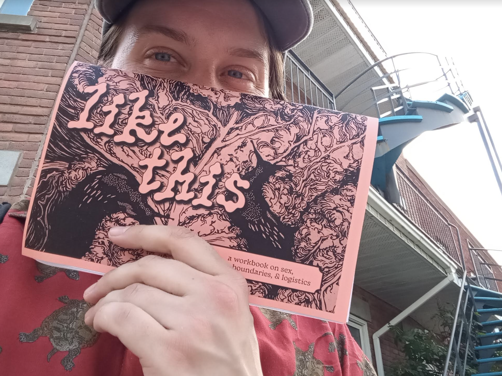Lee, holding up a copy of hir zine "Like This: A Workbook on Consent, Desire, and Logistics" in front of hir face.