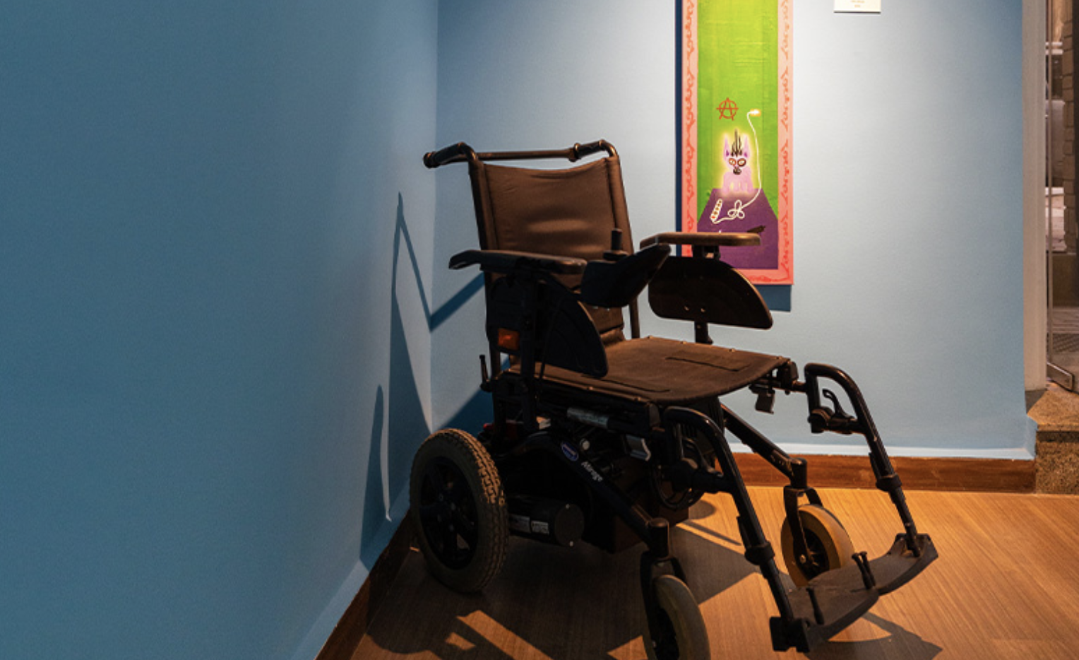 A wheelchair sits in a corner of the "Empty Rooms" exhibit; artwork hanging on wall behind it.