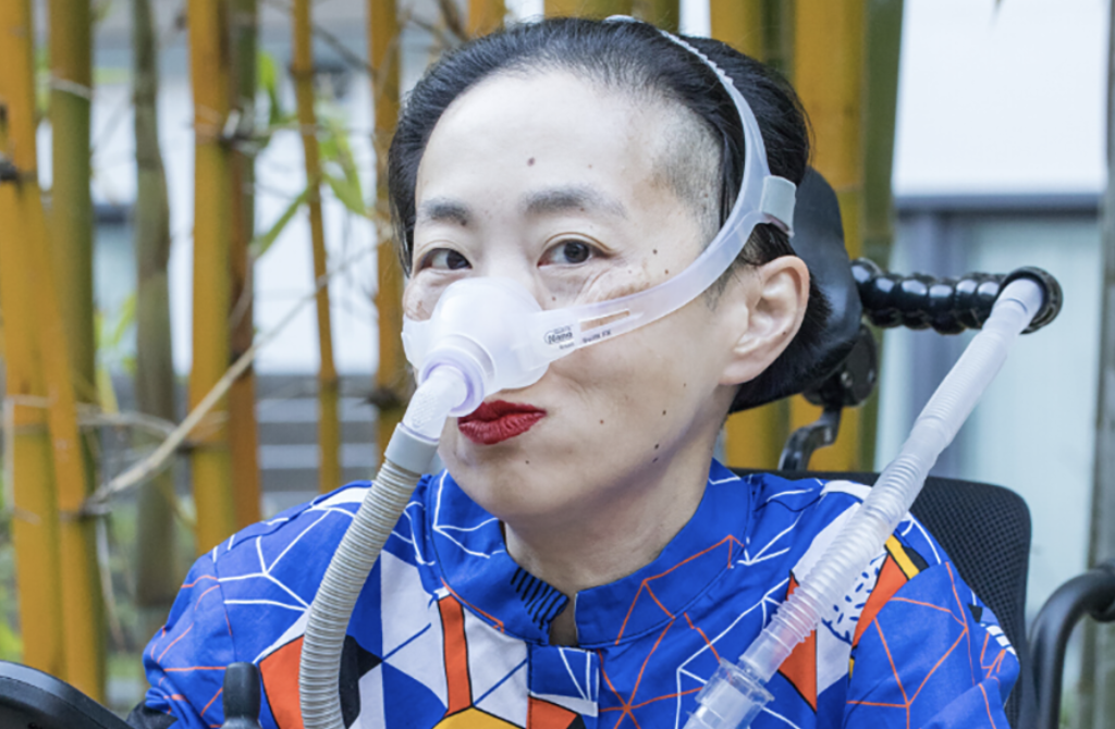 Photo of Alice Wong sitting in her wheelchair, with a mask over her nose to help her breathe. She's wearing red lipstick.