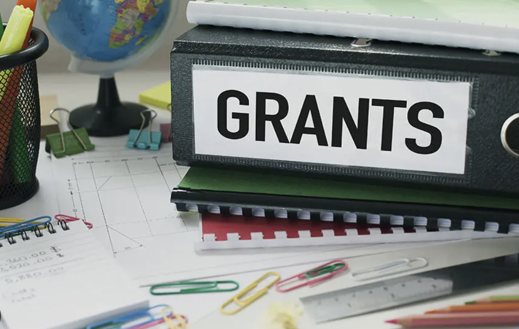 A binder labelled "GRANTS" sits on a desk, stacked on top of other folders.