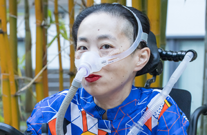 Photo of Alice Wong sitting in her wheelchair, with a mask over her nose to help her breathe. She's wearing red lipstick.